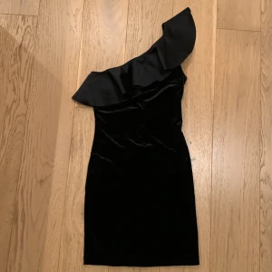Svart one-shoulder klänning från H&M - Säljer en läcker svart one-shoulder klänning från H&M. Klänningen har en snygg volangdetalj över axeln och är gjord i ett glansigt material som ger en lyxig känsla. Perfekt för festliga tillfällen!