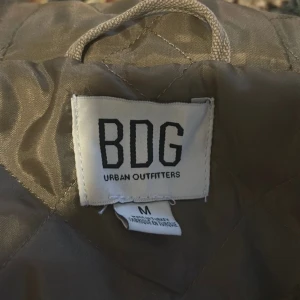 Urban Outfitters  - En snygg jacka med huva från Urban Outfitters. Jackan har en ficka på bröstet och två fickor på sidorna. Den har en dragkedja framtill och en knapp i toppen. Jackan är i gott skick.