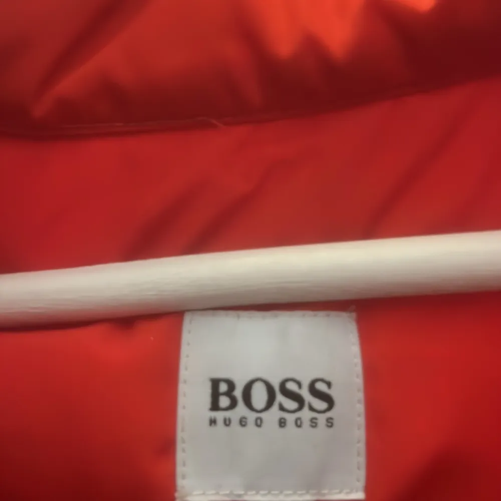 Säljer en snygg röd quiltad väst från Hugo Boss. Västen har en dragkedja framtill och är perfekt för höst- och vårsäsongen. Den är i nyskick och har en stilren design med marinblåa detaljer längs kanterna.. Jackor.