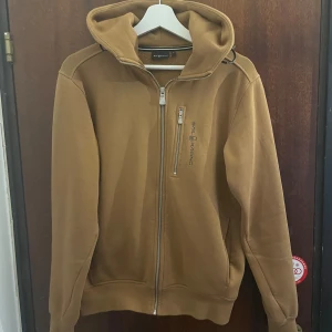 Brun hoodie från Sail Racing - Säljer en snygg brun hoodie från Sail Racing i storlek S. Den har en dragkedja framtill och en bekväm huva. Perfekt för höst och vinter med sin varma och sköna känsla. Hoodien är i bra skick och redo för nya äventyr!