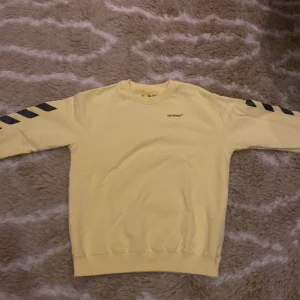 Gul tröja från Off-White - Säljer en supercool gul tröja från Off-White. Den har långa ärmar med svarta diagonala ränder och ett stort svart tryck på ryggen. Perfekt för att sticka ut i mängden! Tröjan är i bra skick och passar perfekt för både vardag och fest.