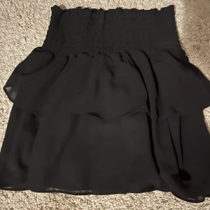 Svart kjol med volangdetaljer - Säljer en superfin svart kjol med volangdetaljer. Den har en elastisk midja som gör den bekväm och lätt att bära. Perfekt för både vardag och fest! från Chelsea jättebra skick🖤