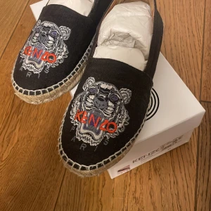 Svarta espadriller från Kenzo - Säljer ett par snygga svarta espadriller från Kenzo med det ikoniska tigerhuvudet broderat i vitt och rött på ovansidan. Skorna är i canvasmaterial och har en klassisk espadrillesstil med flätad sula. 