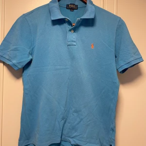Blå pikétröja från Ralph Lauren - Säljer en klassisk blå pikétröja från Ralph Lauren med korta ärmar och den ikoniska loggan broderad i orange på bröstet. Barnstorlek L = vuxenstorlek S