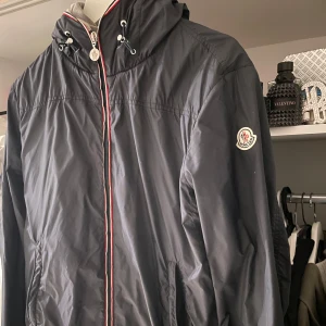 Blå vindjacka från Moncler - Snygg blå vindjacka från Moncler med dragkedja och huva. Jackan har en diskret logga på ärmen och en grå insida som ger en cool kontrast. Perfekt för blåsiga dagar och stilrena outfits. Enda negativa är att ena dragkedjan har tappat själva drag grejen men den funkar fortfarande:) mvh Rhodri 