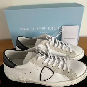 Philippe Model - Snygga vita sneakers med svarta detaljer från Philippe Model. Skorna har en klassisk design med snörning och en stilren look. Perfekta för en trendig och avslappnad stil.