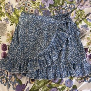 Blå blommig omlottkjol från SHEIN - Charmig blå omlottkjol från SHEIN med ett sött blommönster i vitt. Kjolen har volangdetaljer och knytning i midjan för en feminin touch. Perfekt för en somrig look!