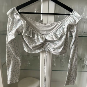 Blommig offshoulder topp med volang - Charmig blommig offshoulder topp med volangdetaljer och knytning framtill. Långärmad design i mjukt material, perfekt för vår och sommar.