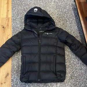 Svart dunjacka från Sail Racing - Säljer en svart dunjacka från Sail Racing med huva och dragkedja. Jackan är perfekt för kyligare väder och har en stilren design med märkets logga på bröstet. Den är både varm och bekväm, idealisk för vintern. Nypris 4500