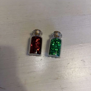 Glitter - Två små glasflaskor fyllda med glitter, en i röd och en i grön färg. Flaskorna har korkar och är perfekta för pyssel eller dekorationer men även för smink och naglar. De glittriga partiklarna ger en festlig känsla och kan användas för att skapa egna smycken eller konstverk. 10kr FÖR BÅDA