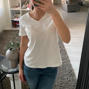 Vit t-shirt med v-ringning  - Säljer en klassisk vit t-shirt från H&M med v-ringning och en bröstficka. Perfekt för en avslappnad stil. T-shirten är kortärmad och gjord i mjuk bomull.