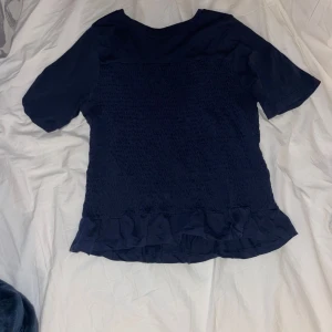 Mörkblå  topp från gina  med volangdetaljer - Säljer en mörkblå topp med korta ärmar och volangdetaljer vid nederkanten. säljer för den e för kort men jätte fin o skön.  Toppen har en smockad textur som ger en snygg passform. Perfekt för att piffa upp vardagslooken!
