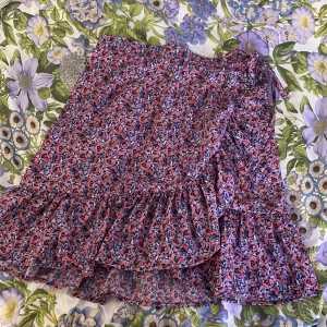 Blommig kjol med volang - Säljer en söt blommig kjol i rosa och blått med volangdetaljer. Kjolen har en omlott-design och knytband i midjan för justerbar passform. Perfekt för sommarens varma dagar!