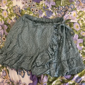 Grön blommig omlottkjol - Charmig grön omlottkjol med blommigt mönster och volangdetaljer. Kjolen har en knytning i sidan och är perfekt för en somrig look. Har ingen lapp men storleken är mellan S/M , Den korta längden och det lätta materialet gör den idealisk för varma dagar.