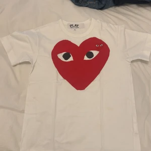 Vit t-shirt med rött hjärta från Comme des Garçons - Säljer en vit t-shirt från Comme des Garçons med ett stort rött hjärta med ögon på framsidan. T-shirten har korta ärmar och en rund halsringning. Perfekt för en stilren och lekfull look.