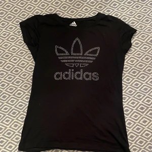 Svart t-shirt från Adidas - Snygg svart t-shirt med rhinestones på från adidas. Bra skick och inte använd så mycket. Den har en mer tajt passform så passar även S och kanske XS. KÖP ENDAST VIA POSTNORD