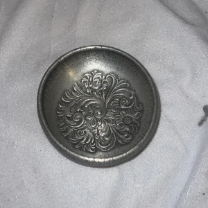 Dekorativ skål i metall - Säljer en vacker dekorativ skål i metall med ett intrikat blommönster på insidan. Skålen har en antik finish och en rund form, perfekt som prydnad eller förvaring av småsaker. En liten nyckel medföljer.