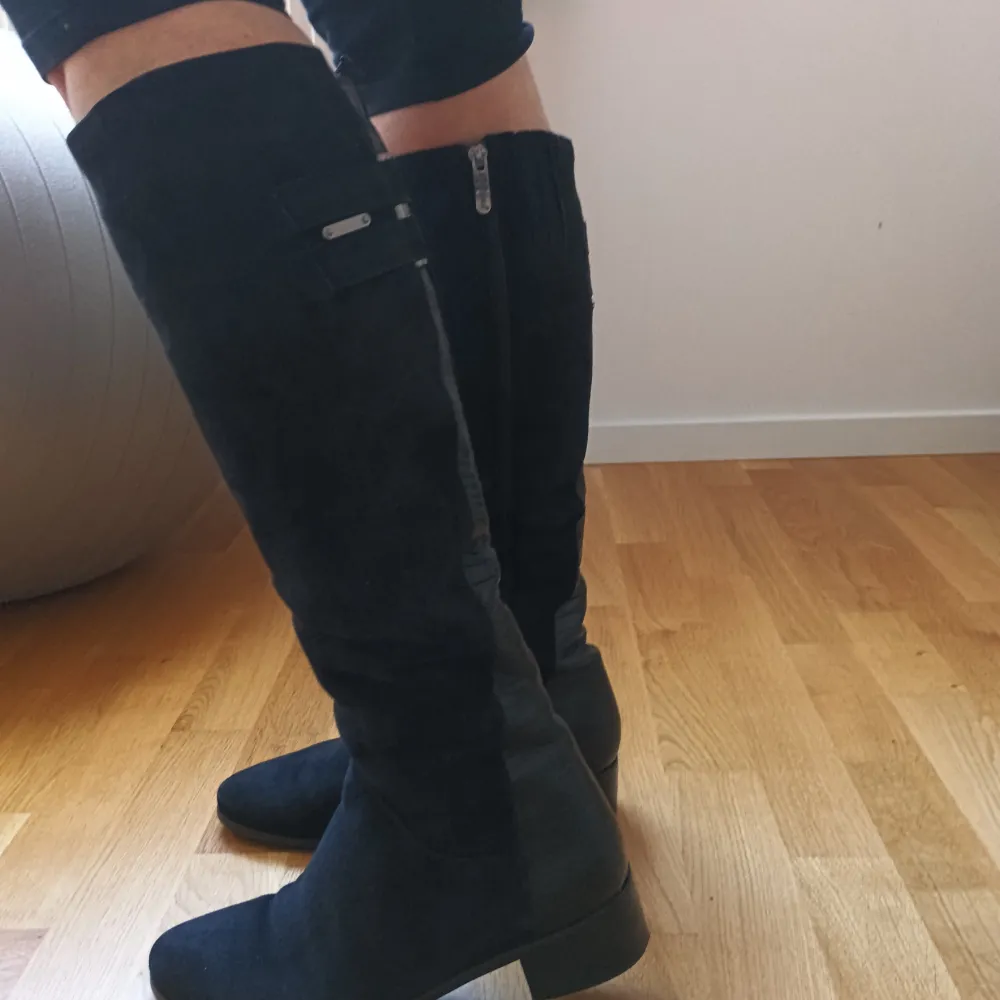 Snygga svarta stövlar i mocka med krokodilmönstrade detaljer och dragkedja på sidan.vinterstövlar i äkta läder med äkta päls inuti. Perfekta för en stilren look. De har en låg klack och är knähöga, vilket ger en elegant touch. . Kengät.