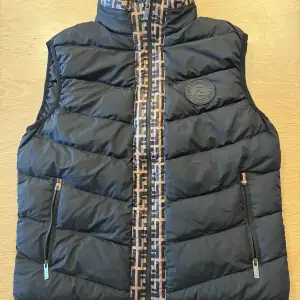 Snygg svart dunväst från Fendi med ett stilrent mönster i brunt längs dragkedjan. Västen har två praktiska fickor med dragkedjor och ett Fendi-märke på bröstet. 1.1k