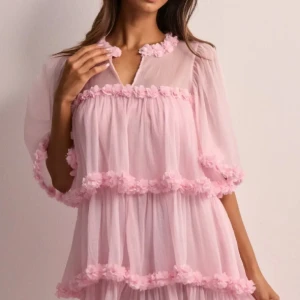 Rosa klänning med blommiga detaljer - Söt rosa klänning med blommiga volangdetaljer och transparenta trekvartsärmar. Perfekt för en romantisk look med sin luftiga design och mjuka färgton.