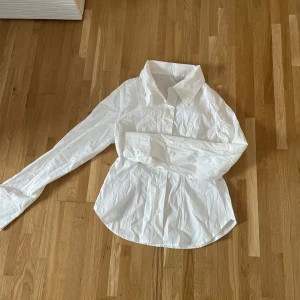 Vit skjorta med knappar - Säljer en klassisk vit skjorta med långa ärmar och knappar framtill. Perfekt för en stilren look. Skjortan har en enkel design med en krage som ger en tidlös känsla.