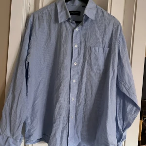 Blå rutig skjorta från Giorgio Armani - Säljer en stilren blå rutig skjorta från Giorgio Armani Collezioni. Skjortan har långa ärmar och knappar framtill. Perfekt för en elegant look.