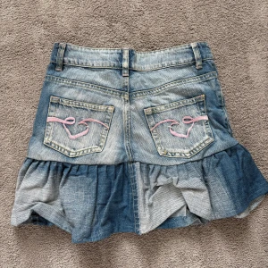 Jeanskjol med volangdetaljer från Check His - Snygg jeanskjol från Check His med volangdetaljer och rosa broderi på bakfickorna. Kjolen har en klassisk jeanslook med bälteshällor och dragkedja framtill. Perfekt för en avslappnad stil med en twist.