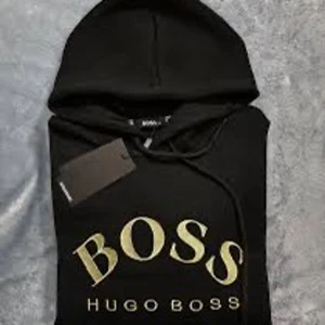 Svart hoodie från Hugo Boss - Snygg svart hoodie från Hugo Boss med guldtryck på bröstet. Den har en klassisk design med huva och dragsko samt en praktisk magficka. Perfekt för en stilren och bekväm look. Pris kan diskuteras