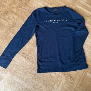 Mörkblå långärmad tröja från Tommy Hilfiger - Säljer en stilren mörkblå långärmad tröja från Tommy Hilfiger i ekologisk bomull. Tröjan har en klassisk rund halsringning och logotypen tryckt på bröstet. Perfekt för en avslappnad och trendig look. 🌟
