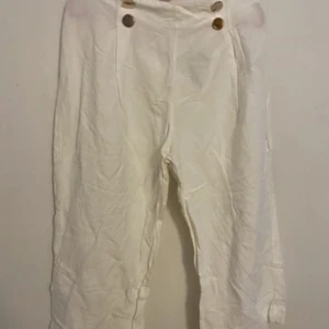 Vita byxor från Holly & Whyte - Snygga vita byxor från Holly & Whyte by Lindex med hög midja och dekorativa knappar framtill. Perfekta för en stilren look. Tillverkade i ett bekvämt material som passar bra till olika tillfällen.