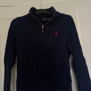 Mörkblå stickad halfip tröja frånRalph Lauren - Snygg mörkblå stickad tröja från Polo Ralph Lauren med dragkedja vid halsen och det ikoniska röda logotypbroderiet på bröstet. Perfekt för kyligare dagar med sin kabelstickade design.