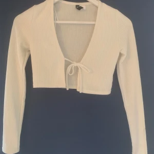Vit ribbad kofta från H&M Divided - Säljer en stilren vit ribbad kofta från H&M Divided. Den har en croppad design med långa ärmar och knytband framtill. Perfekt för lager-på-lager-stil eller som ett lättare plagg under svalare dagar. Har fläck på ärmen. 