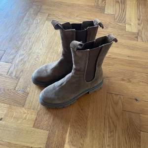 Bruna mocka boots - Snygga bruna boots i mocka med grov sula och elastiska paneler på sidorna för enkel på- och avtagning. Perfekta för höst och vinter med en robust design som ger bra grepp. Storlek 39.