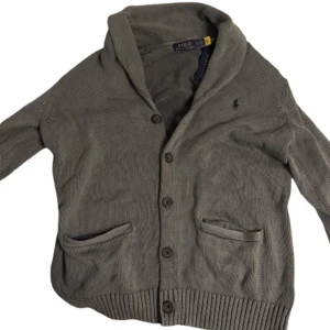 Grå kofta från Polo Ralph Lauren - Säljer en stilren grå kofta från Polo Ralph Lauren med knappar framtill och två fickor. Koftan har en klassisk krage och är perfekt för kyligare dagar. Den har långa ärmar och ribbade muddar för en bekväm passform. Storlek s men passar m