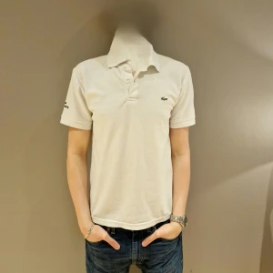 Vit pikétröja från Lacoste - Säljer en klassisk vit pikétröja från Lacoste med korta ärmar och den ikoniska krokodilloggan på bröstet. Perfekt för en stilren och avslappnad look. Tröjan har en krage och knappar framtill.