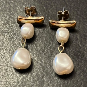 Guldörhängen med vita pärlor - Eleganta örhängen i guld med två vita pärlor i varje. Den översta delen är en guldstång som fäster i örat, och under hänger två oregelbundna vita pärlor. Perfekt för att ge en lyxig touch till din stil.