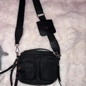Svart axelväska med justerbar rem - Snygg svart axelväska med flera dragkedjefickor och justerbar axelrem. Väskan har en praktisk design med extra ficka på remmen och detaljer i metall. Perfekt för att hålla ordning på dina saker.