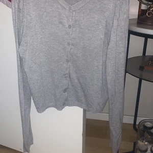 Grå kofta med knappar - Säljer en stilren grå kofta med knappar framtill. Den har en v-ringad design och långa ärmar, perfekt för lager på lager under kyligare dagar. Koftan är i ett mjukt material som ger en bekväm passform. Storlek L men passar för S/M