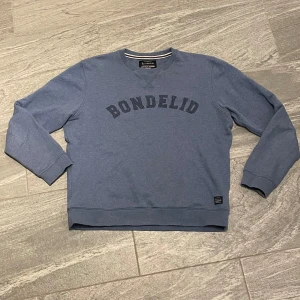 Blå sweatshirt från Bondelid - Säljer en stilren blå sweatshirt från Bondelid med tryckt logotyp på bröstet. Tröjan har långa ärmar och ribbade muddar vid ärmslut och nederkant. Perfekt för en avslappnad look.