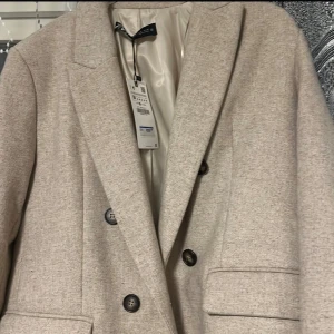 Beige kappa från Zara - Elegant beige kappa från Zara med klassisk design och knappar framtill. Perfekt för en stilren look. Kappan har en mjuk och bekväm passform med fickor på sidorna.