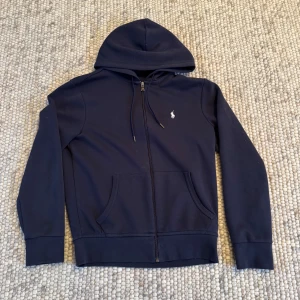 Mörkblå hoodie från Polo Ralph Lauren - Säljer en stilren mörkblå hoodie från Polo Ralph Lauren med dragkedja och klassisk logga på bröstet. Tröjan har långa ärmar och en praktisk ficka framtill. Perfekt för en avslappnad look.