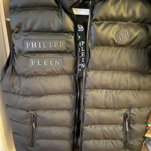 Svart dunväst från Philipp Plein - Säljer en svart dunväst från Philipp Plein med dragkedja och logotypdetaljer på bröstet. 