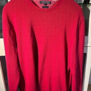 Röd tröja från Tommy Hilfiger - Säljer en snygg röd tröja från Tommy Hilfiger i storlek XL. Tröjan har en klassisk design med rund hals och långa ärmar. Perfekt för en stilren look. Har dock två väldigt små nästintill omgöligt att se fläckar på ena armen, se bild