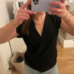 Svart omlottopp från Zara - Snygg svart omlottopp från Zara med korta ärmar och v-ringning. Perfekt för en stilren look. Passar bra till både jeans och kjol.