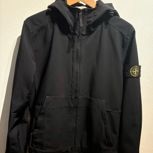 Svart jacka med huva från Stone Island - Snygg svart jacka med huva från Stone Island. Jackan har dragkedja framtill och en ikonisk logotyp på ärmen. Perfekt för en stilren look.
