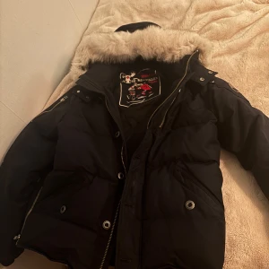 Svart parkajacka från Moose Knuckles - Säljer en svart parkajacka från Moose Knuckles med pälskantad huva och dragkedja. Jackan har flera fickor och en broderad logga på insidan.
