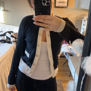 Svart kofta med vita detaljer - Snygg svart kofta med guldiga knappar och detaljer. Perfekt för lager-på-lager-stil. Koftan har långa ärmar och en klassisk rund halsringning. Köpt på Plick men nästan aldrig använd