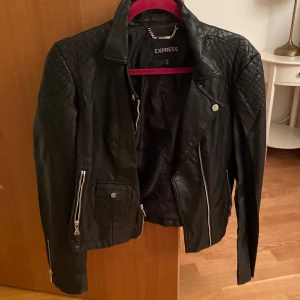 Svart skinnjacka från Express - Snygg svart skinnjacka från Express. Använd och lite sliten vid kragen men syns ej när man har på sig den. 