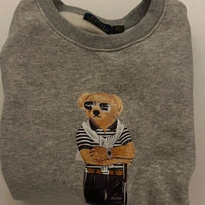 Grå sweatshirt från Ralph Lauren - Säljer en grå sweatshirt från Ralph Lauren med en cool Polo Bear-design på framsidan. Tröjan har långa ärmar och rund halsringning, perfekt för en avslappnad stil. 🐻