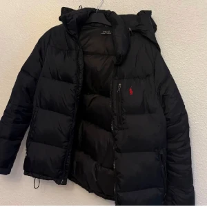 Svart dunjacka från Polo Ralph Lauren - Snygg svart dunjacka från Polo Ralph Lauren med huva och dragkedja. Jackan har en diskret logga på bröstet och är perfekt för kyligare väder. Ny pris 6800